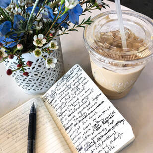 Handwritten poem in a notebook surrounded by a vase of flowers and an iced coffee.Picture